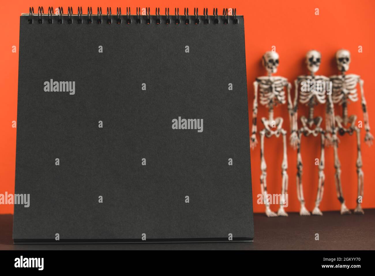 Notebook nero vuoto su sfondo arancione con scheletri sfocati. Design minimalista per le feste di Halloween. Mock up. Copia spase. Foto Stock