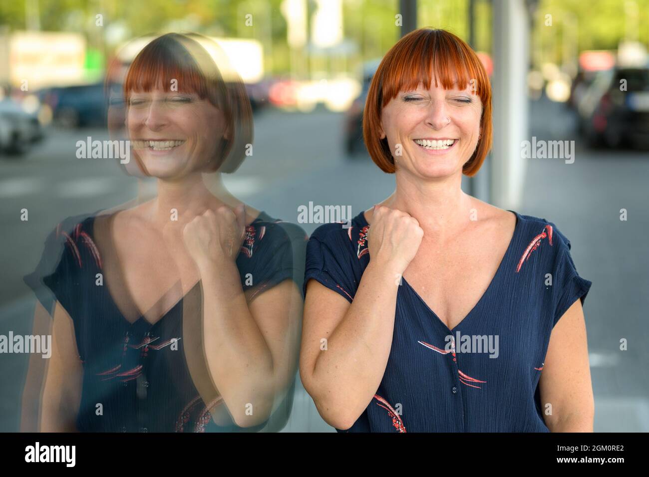 Eccitata donna rossa di mezza età trionfante che pompa l'aria con il suo pugno con un sorriso raggiante mentre lei si leva in piedi contro una grande finestra in città r Foto Stock