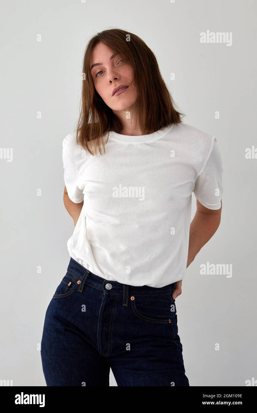 Modello femminile positivo indossando maglietta bianca e jeans in piedi con le mani sulla vita su sfondo bianco e guardando la fotocamera Foto Stock