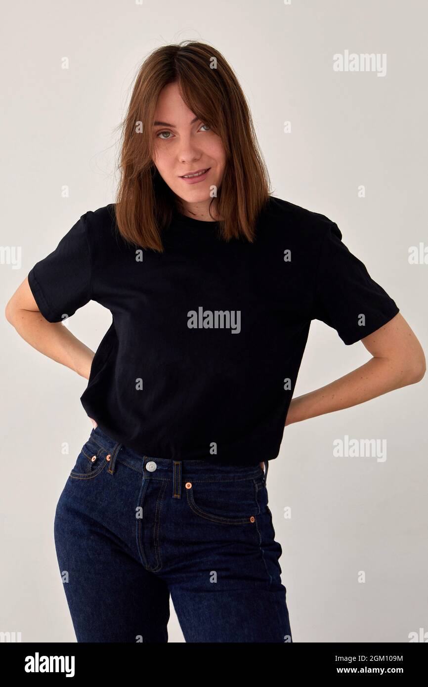 Modello femminile positivo indossando maglietta nera e jeans in piedi con le mani sulla vita su sfondo bianco e guardando la fotocamera Foto Stock