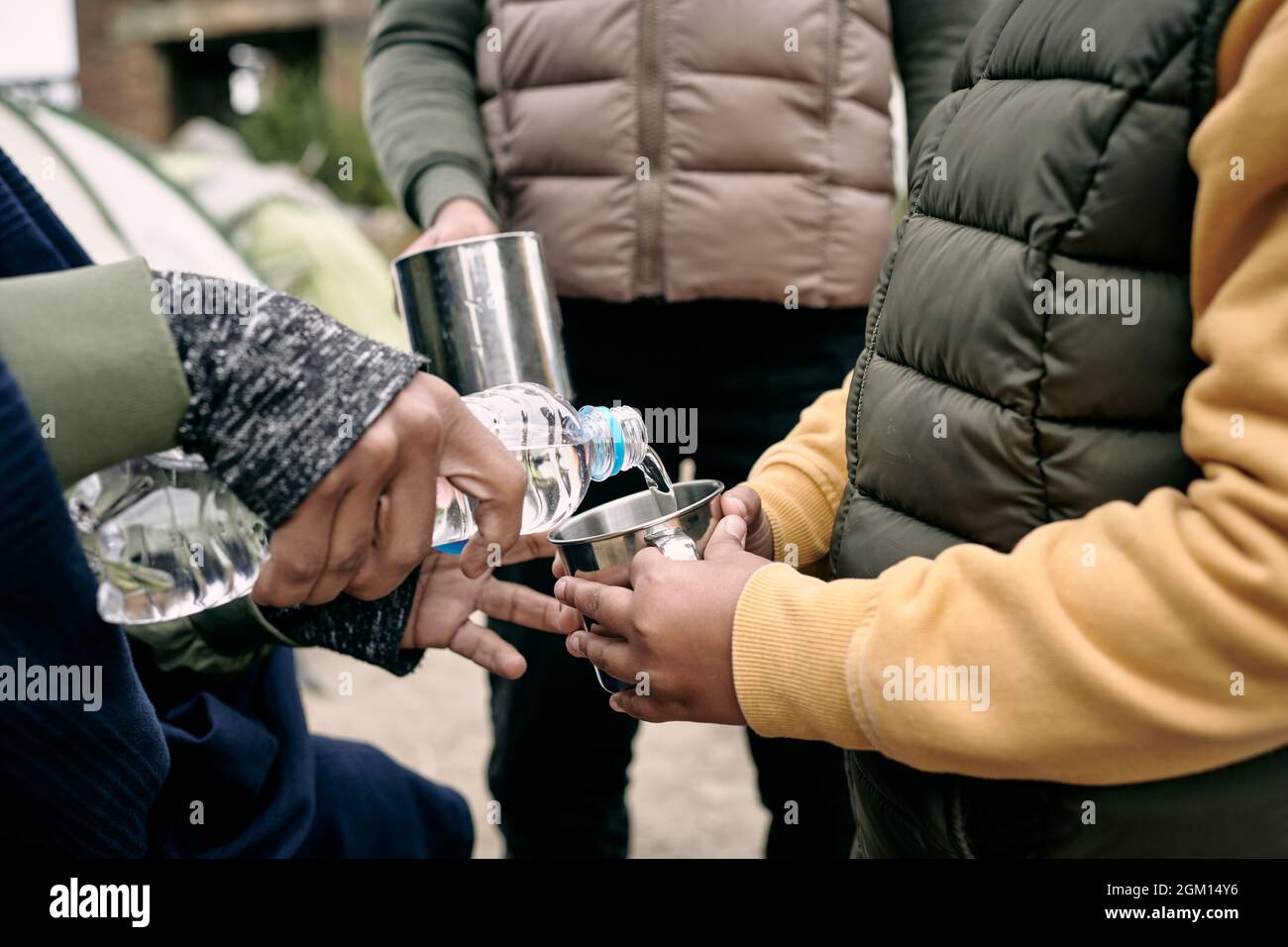 Primo piano di una madre irriconoscibile che versa acqua in tazze metalliche ai bambini mentre vivono nel campo profughi Foto Stock