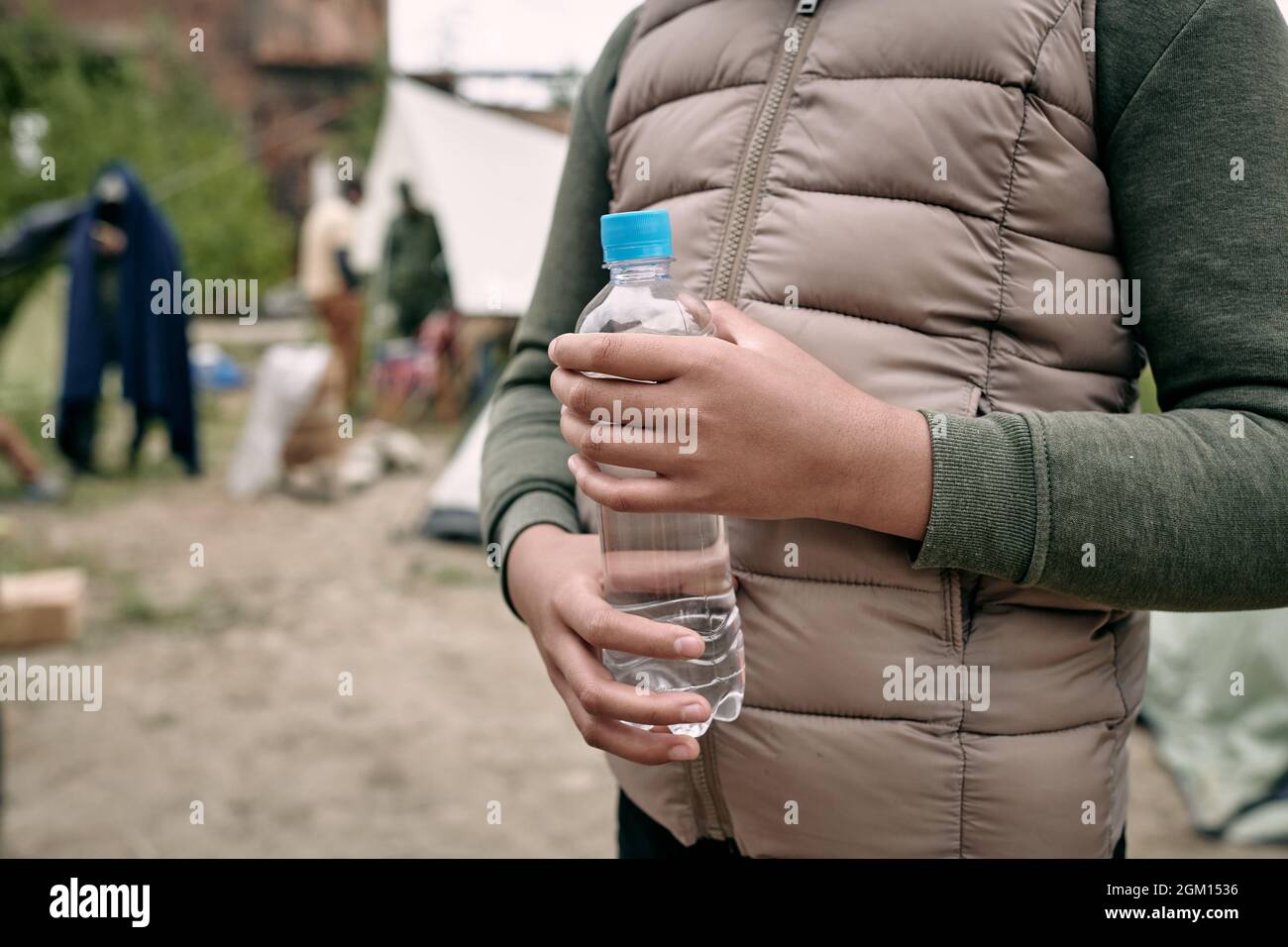 Primo piano di una ragazza nera irriconoscibile in un caldo gilet in piedi con una bottiglia d'acqua contro il campo migrante Foto Stock
