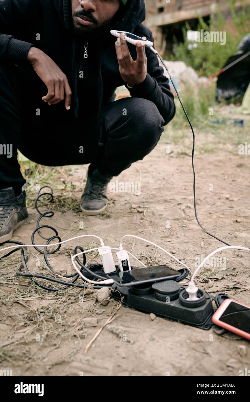 Primo piano di un uomo rifugiato nero accoccolato alla presa di corrente e che ascolta il messaggio audio sul telefono durante la ricarica all'aperto Foto Stock