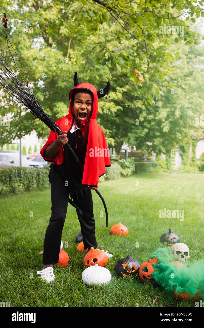 spaventoso ragazzo afroamericano in costume diavolo di halloween che tiene scopa vicino zucche sul prato Foto stock Alamy