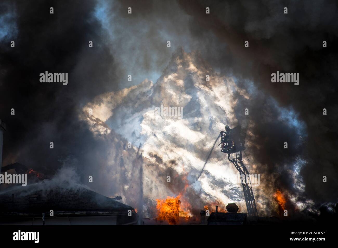 I vigili del fuoco volontari fanno coniare l'acqua da una gru su un tetto che brucia dall'alto. Si staglia contro la montagna innevata e incorniciata da fiamme e fumo. Foto Stock