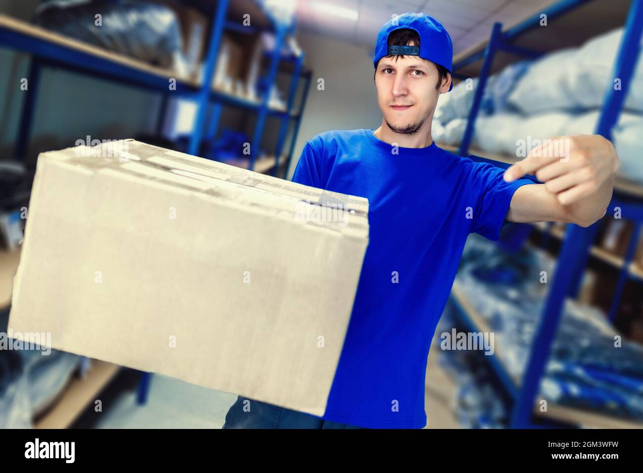 Un addetto ai servizi di consegna tiene una scatola in mano contro lo sfondo dei rack del magazzino. Il corriere in tute blu punta al pacco. Layout con spazio da copiare. Foto Stock
