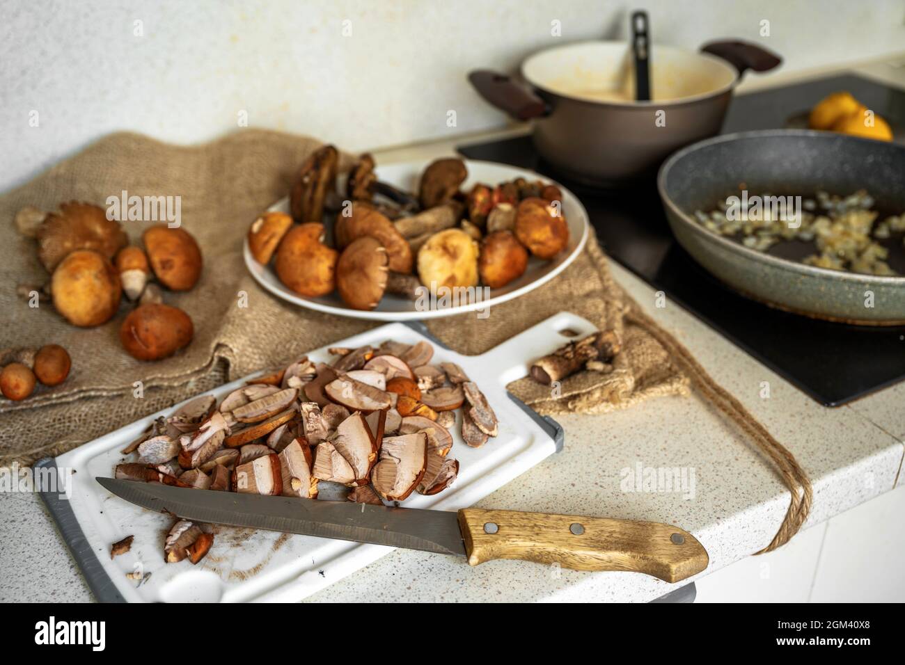 Tavolo da cucina con molti funghi forestali, interi e tagliati a fette con coltello. Preparazione di salsa di funghi su stufa. Foto Stock