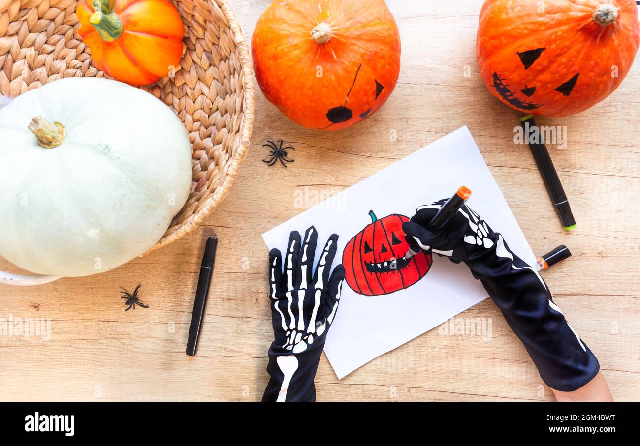 Su una scrivania di legno circondata da zucche e ragni, le mani in guanti neri con ossa stanno disegnando uno schizzo con marcatori una zucca ridente, vista dall'alto Foto Stock