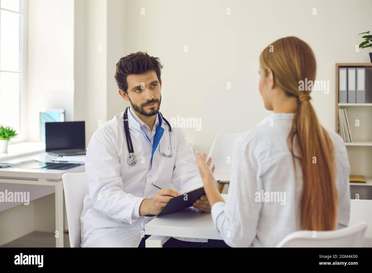 Un attento medico maschile in ospedale consulta la paziente femminile e registra i disturbi della salute. Foto Stock