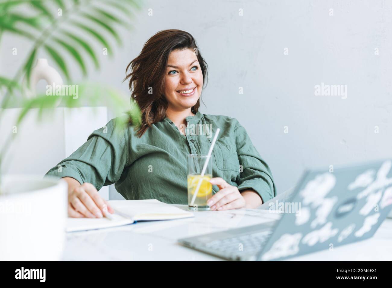 Giovane donna bruna sorridente più taglia in camicia casual con acqua di limone che lavora al computer portatile nel luminoso ufficio moderno Foto Stock