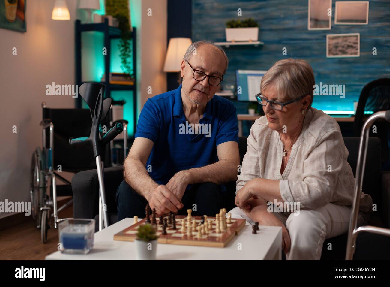 Gli anziani godono di pensione con gioco di scacchi sul tavolino da caffè in soggiorno. Coppia anziana seduta sul divano con scacchiera che gioca insieme per divertimento e divertimento a casa Foto Stock