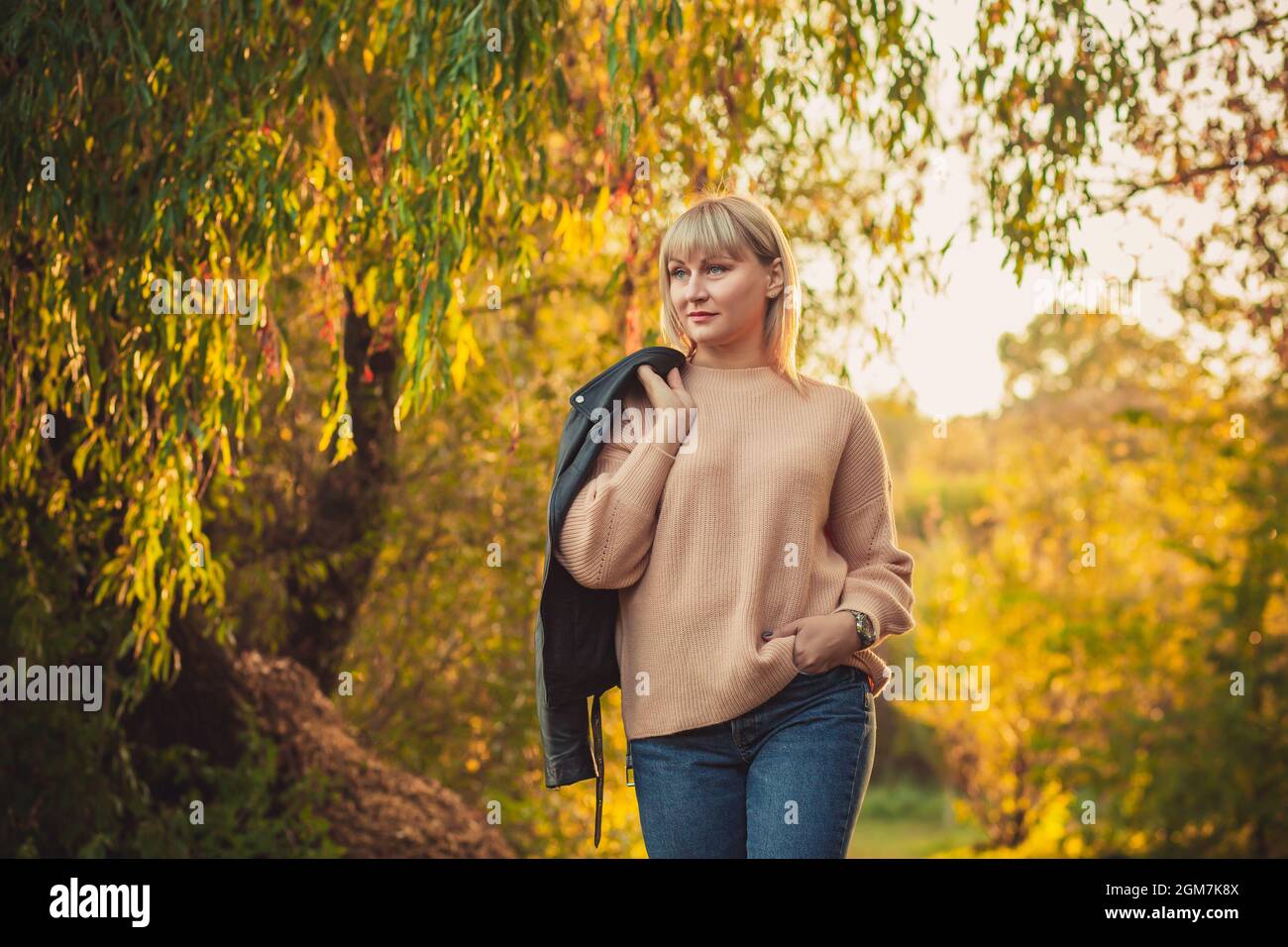 Una donna bionda con un taglio corto cammina attraverso la foresta in un maglione a maglia e una giacca in pelle sulla spalla. Escursioni all'aperto in autunno fores Foto Stock