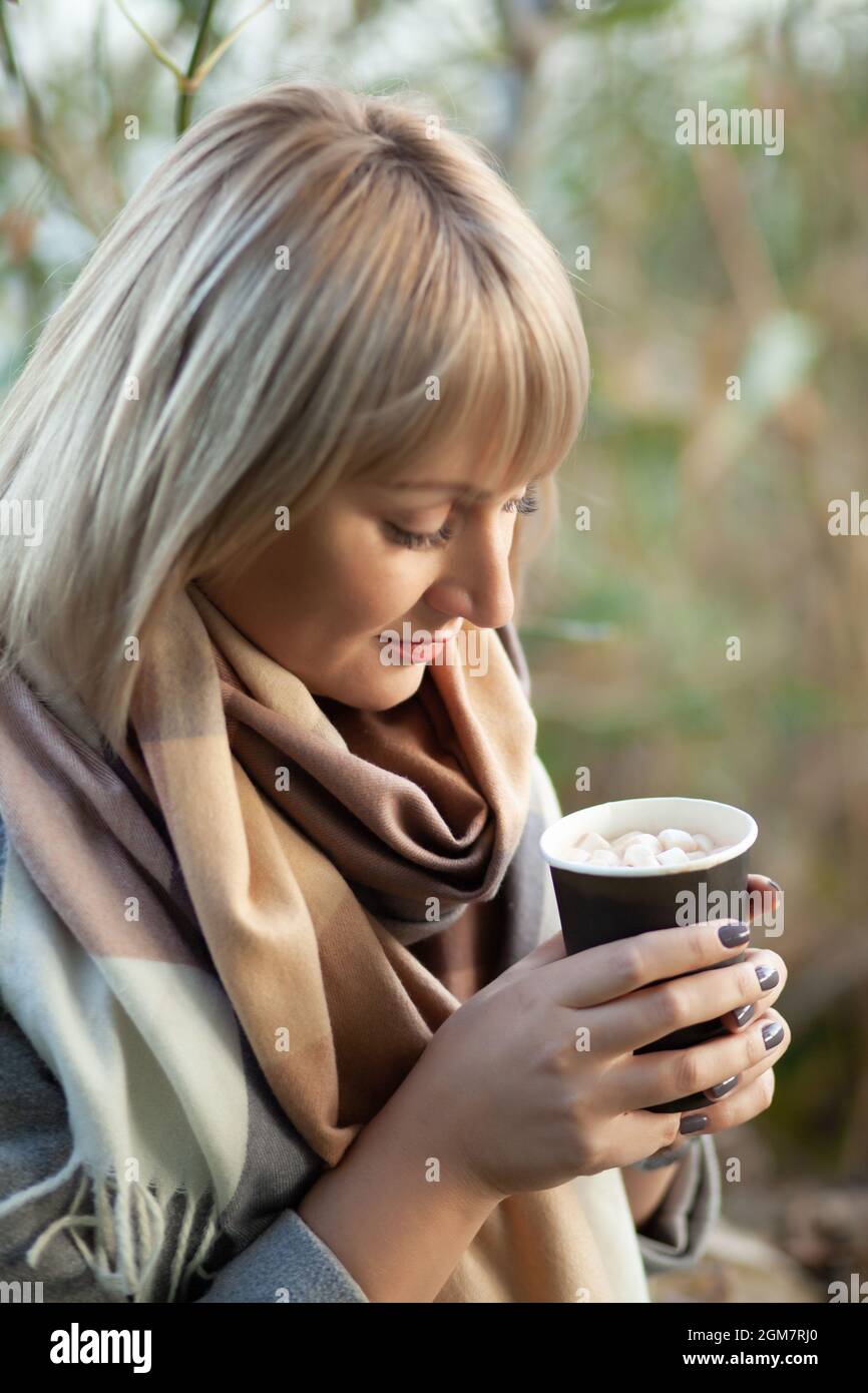 Donna bionda con taglio corto in lana di colore grigio e sciarpa a pipa beve cappuccino al caffè con marshmallows. Passeggiate e pic-nic in natura in autunno f Foto Stock