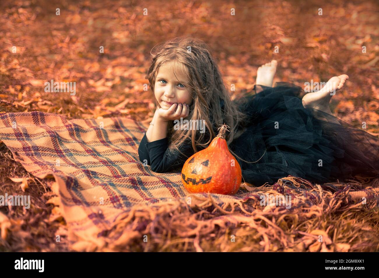 Halloween. Una ragazza carina con capelli lunghi si trova su una coperta arancione nel parco accanto a una zucca divertente. Foto Stock