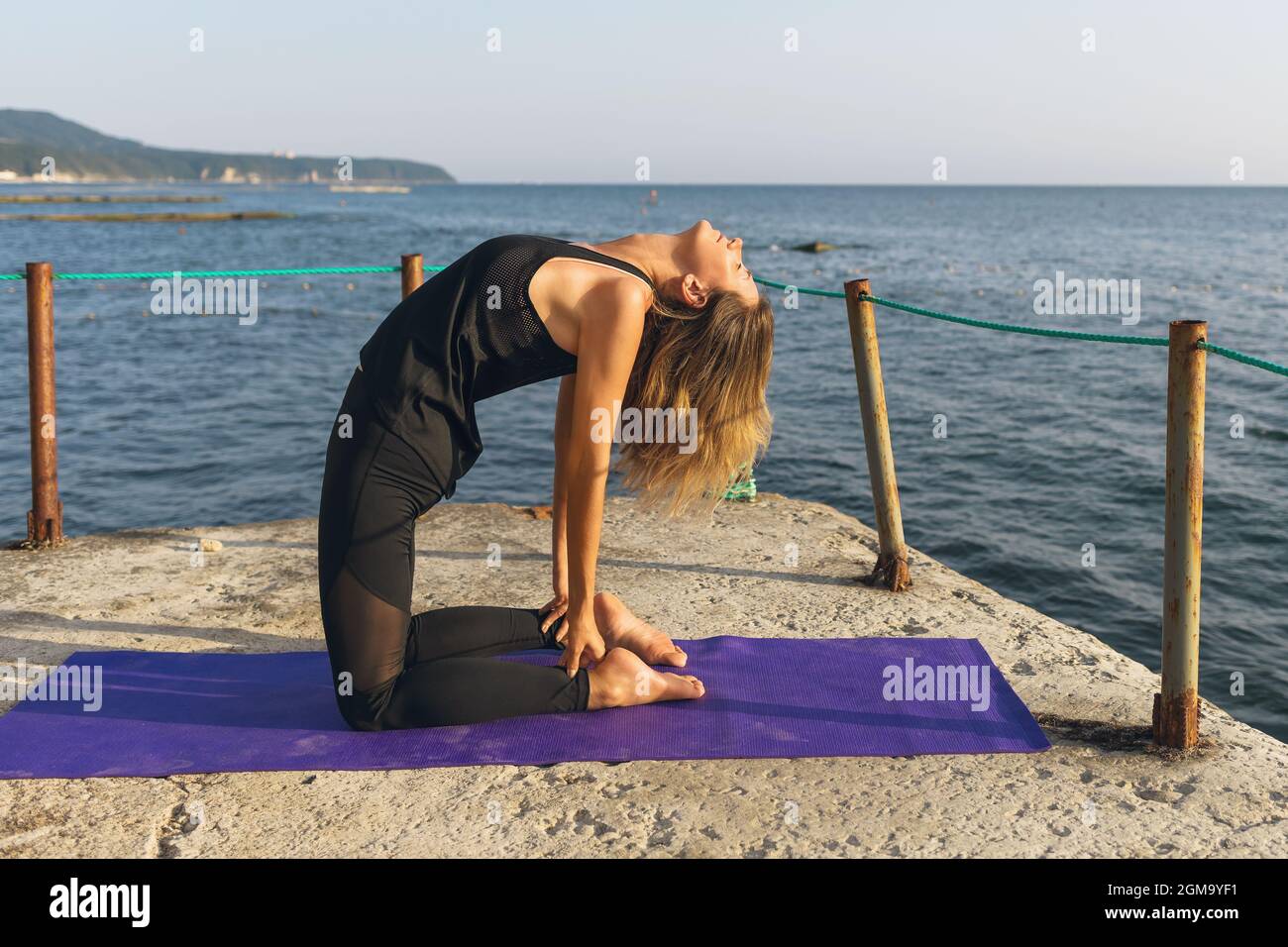 Una donna in abbigliamento sportivo, pratica yoga, su un molo in una soleggiata serata estiva, esegue l'esercizio di hushtrasana, posa cammello Foto Stock