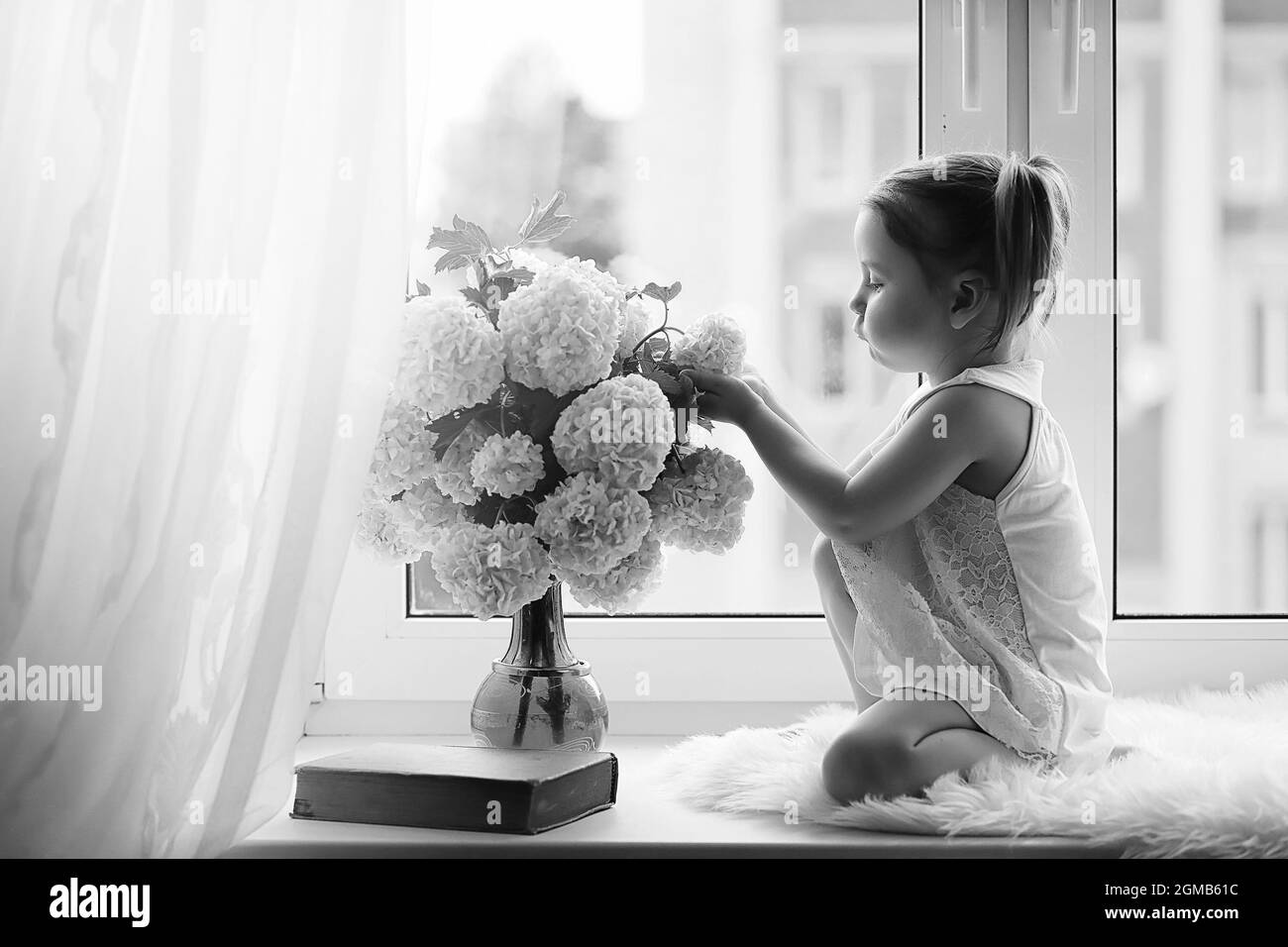 La bambina è seduta sul davanzale. Un mazzo di fiori in un vaso dalla finestra e una ragazza lo sniffing fiori. Una piccola principessa in un bianco dre Foto Stock