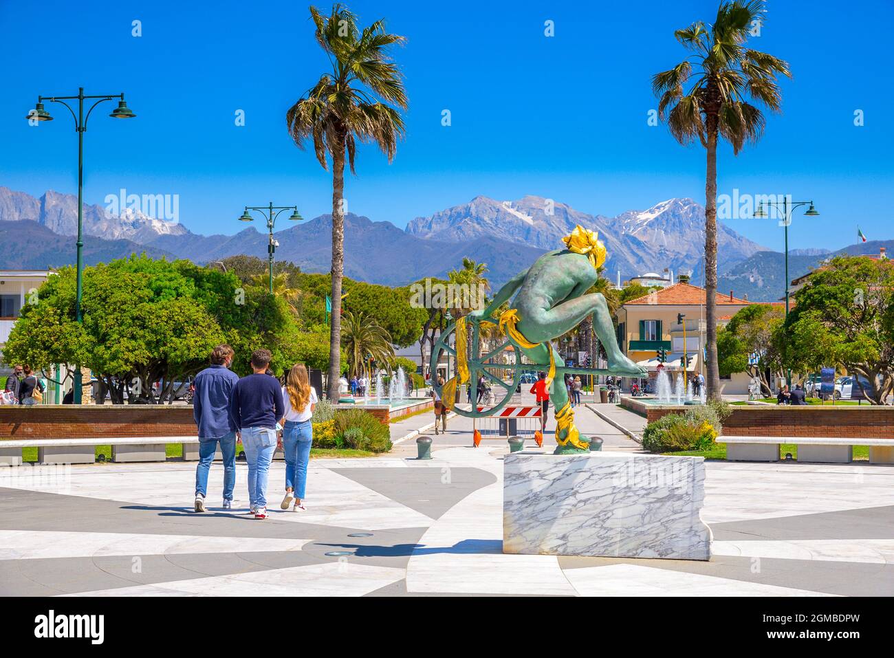 la gente cammina nella piazza, il pavimento in marmo, una scultura dorata in bronzo nella piazza centrale. Passeggiata sul molo. Apuane montagne sullo sfondo, palme Foto Stock