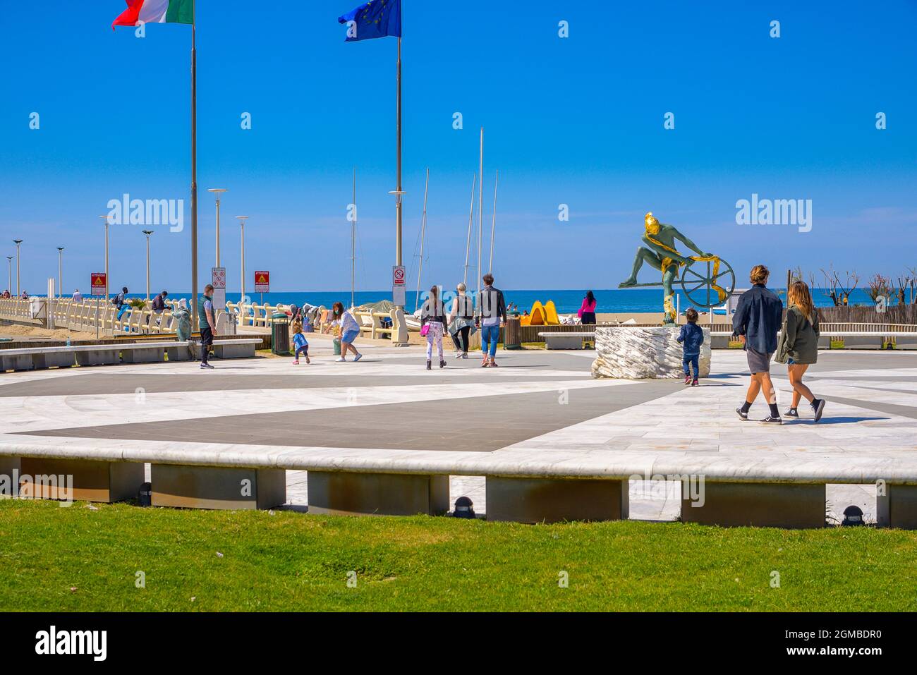 la gente cammina nella piazza con un pavimento in marmo, una scultura in bronzo e oro nella piazza centrale. Promenade sul molo, spiaggia di mare blu. Italiano e bandiere Foto Stock