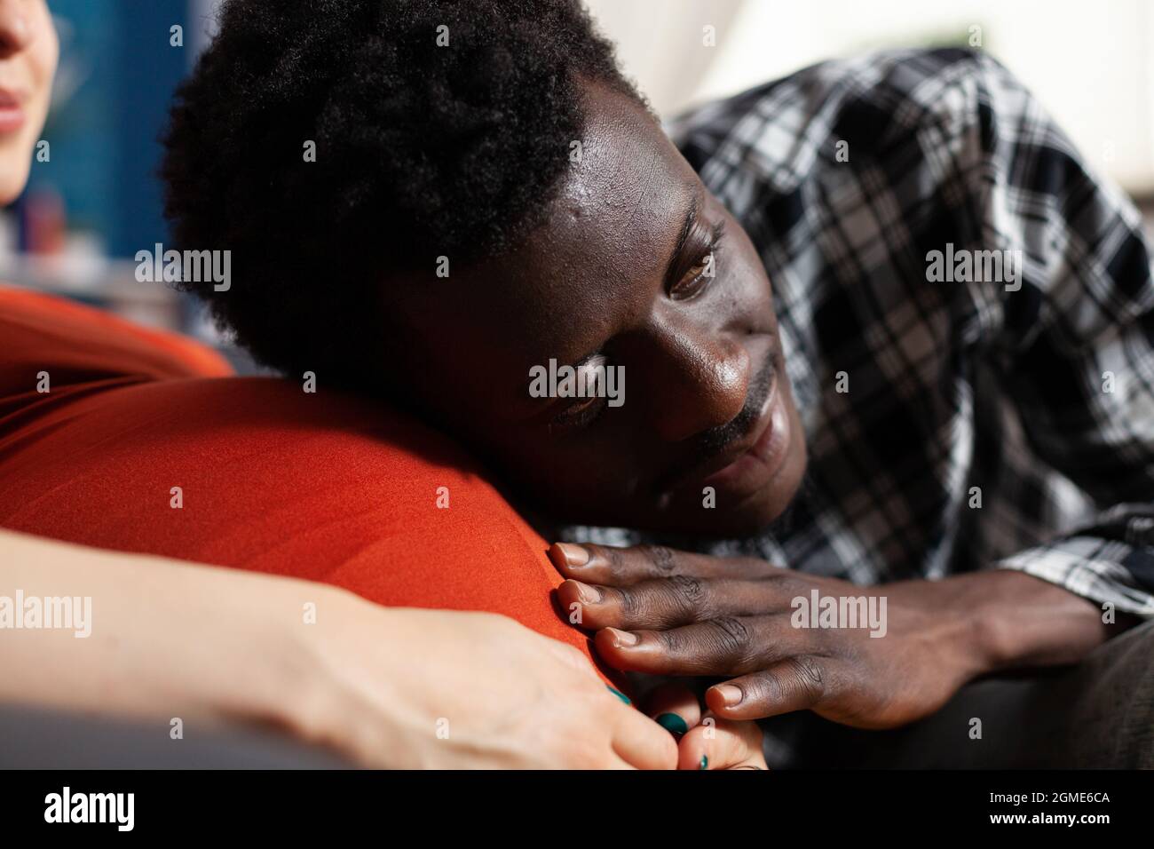 Primo piano di uomo nero che mette la testa sul ventre della donna incinta. Uomo afroamericano che tiene la mano e tocca il baby bump sorridendo. Coppia interrazziale con gravidanza attesa bambino. Foto Stock