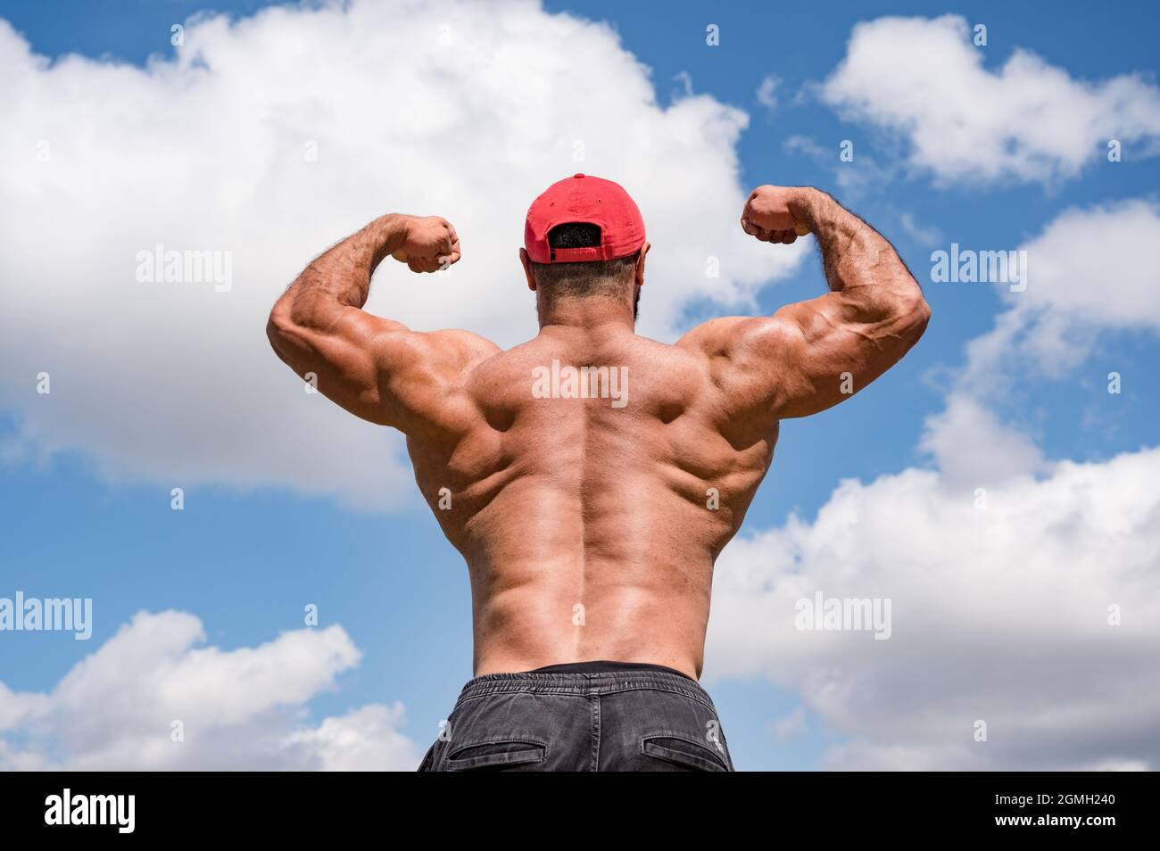 forte giovane atleta fitness bodybuilder maschio in berretto rosso mostrando il suo potente sviluppo muscolare schiena su sfondo cielo blu nuvoloso Foto Stock