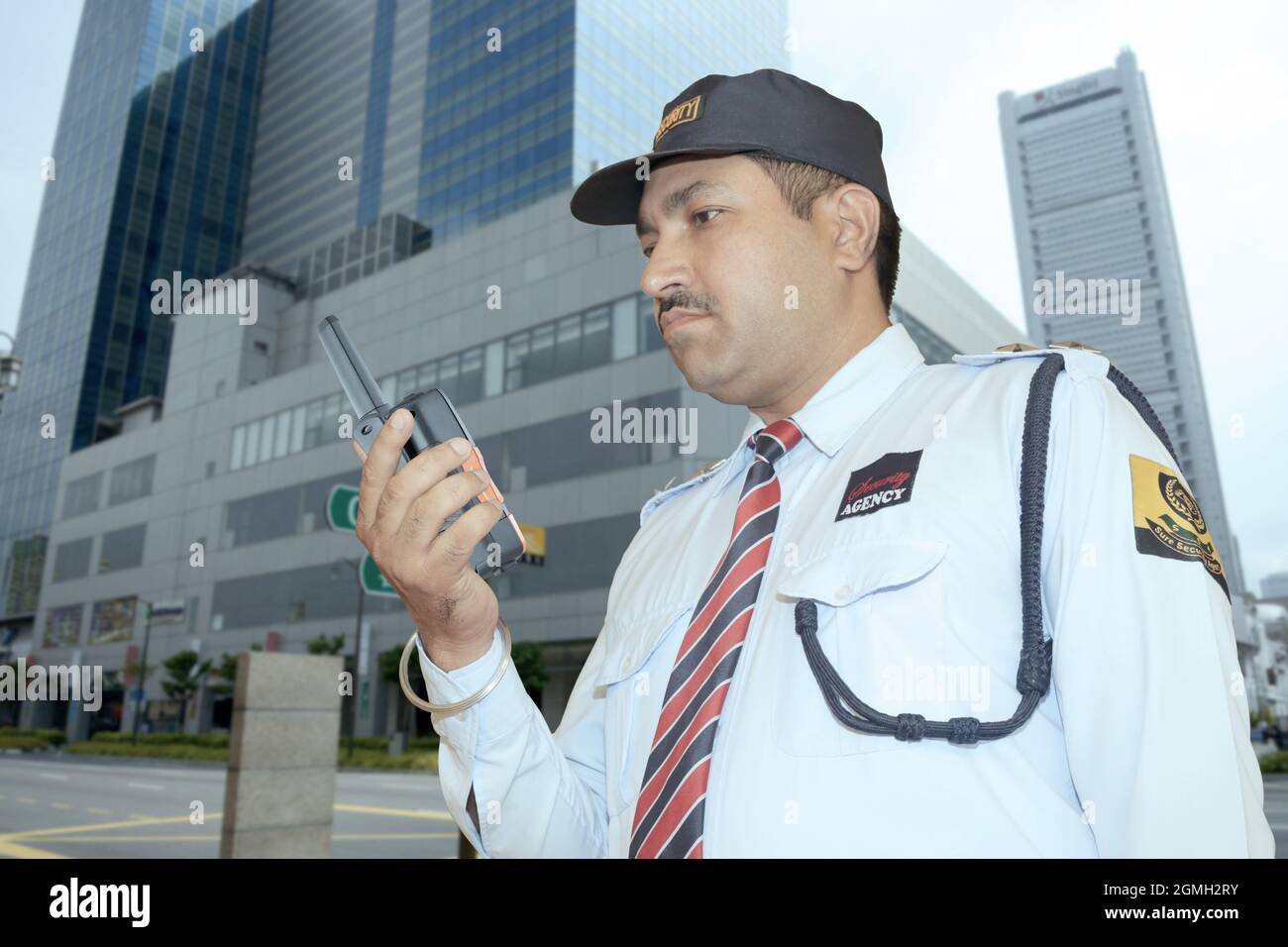 Addetto alla sicurezza in un edificio esterno in servizio con walkie talkie in mano in abito blu con berretto Foto Stock