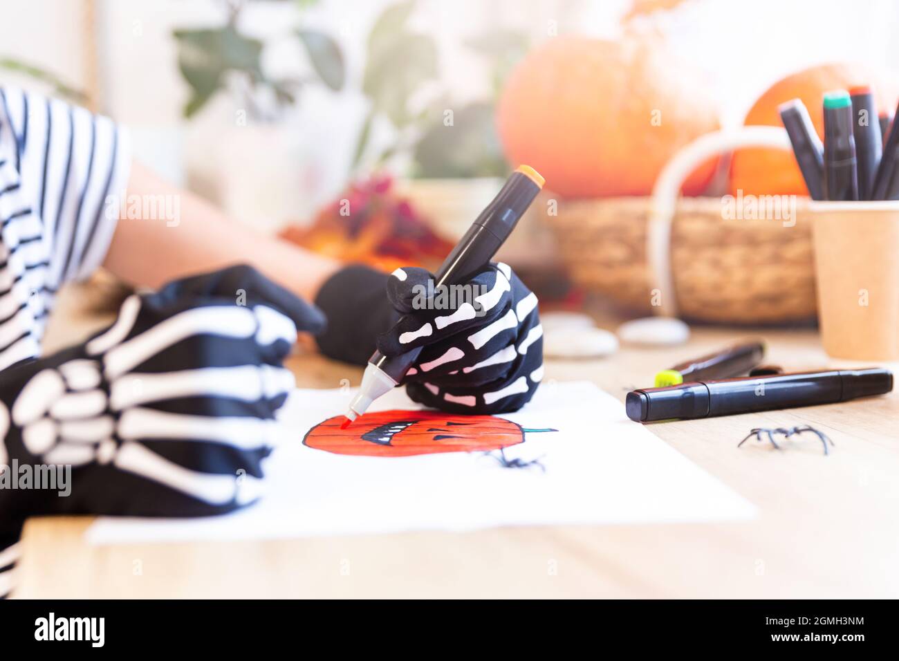 Un bambino disegna con la mano sinistra in guanti neri con ossa una zucca ridente per Halloween, seduta dalla finestra ad una scrivania, zucche in un cestino dentro Foto Stock