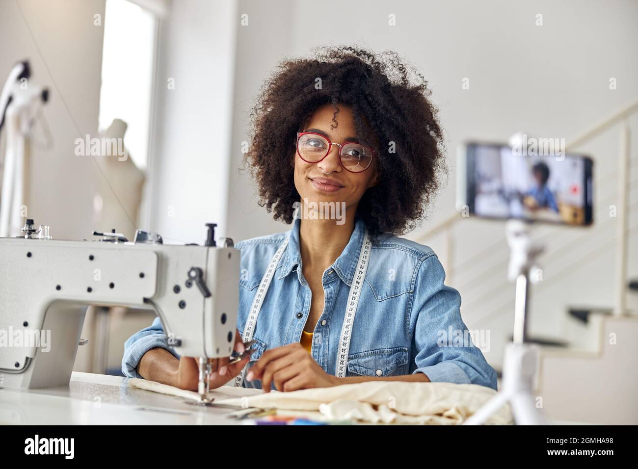 Bella donna afro-americana blogger con forbici e occhiali sul posto di lavoro in laboratorio di cucito Foto Stock