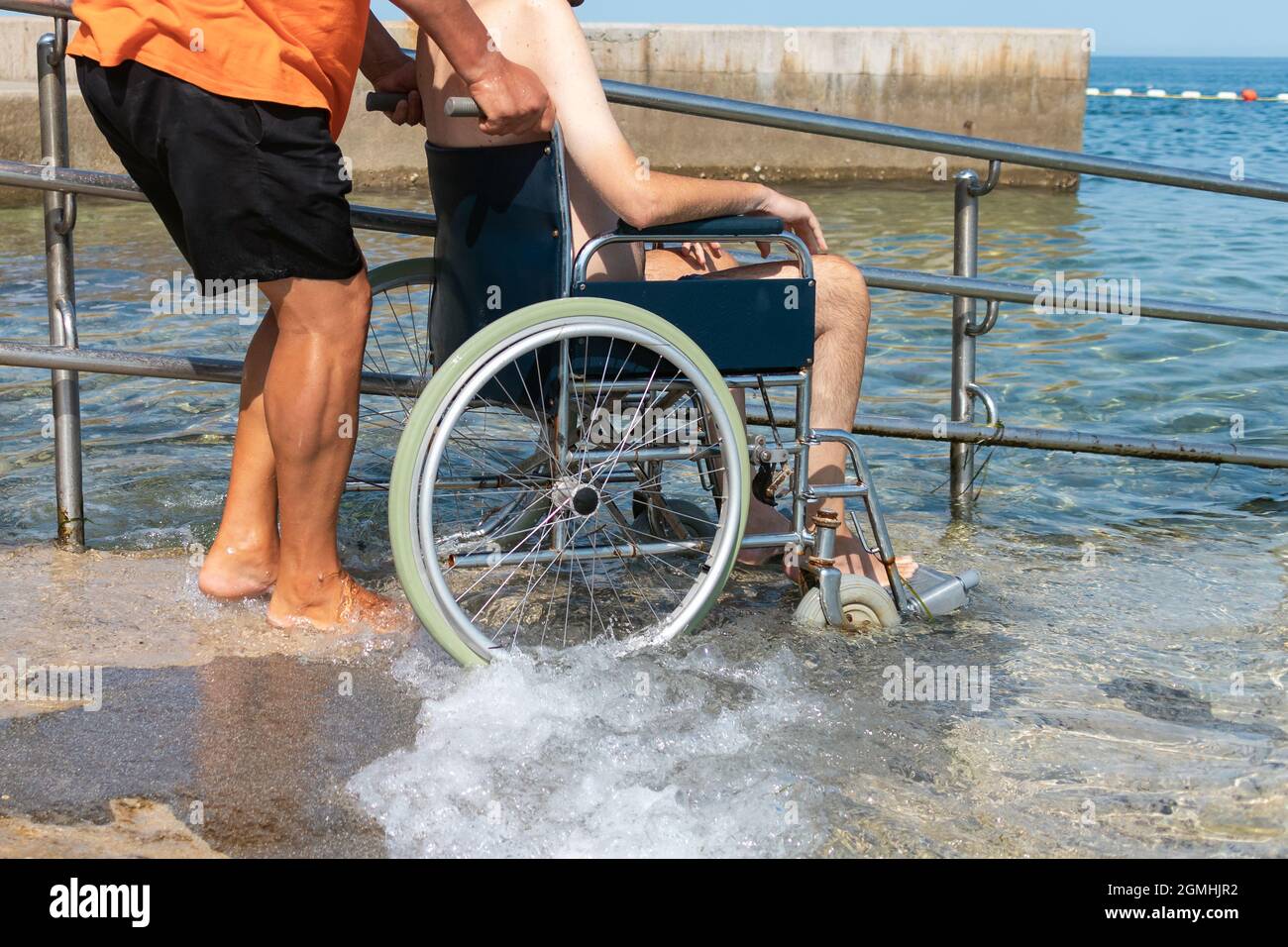 L'uomo spinge lentamente e con attenzione la persona disabile seduta in sedia a rotelle, lungo la rampa sulla spiaggia verso il mare Foto Stock