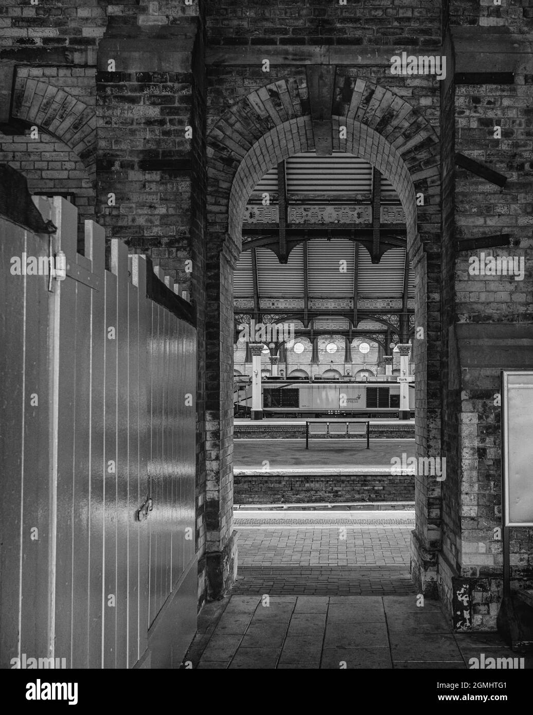 Una piattaforma della stazione ferroviaria e una locomotiva possono essere viste attraverso un arco. Su un lato è presente una partizione bianca in legno. La luce del sole cade sul plat Foto Stock
