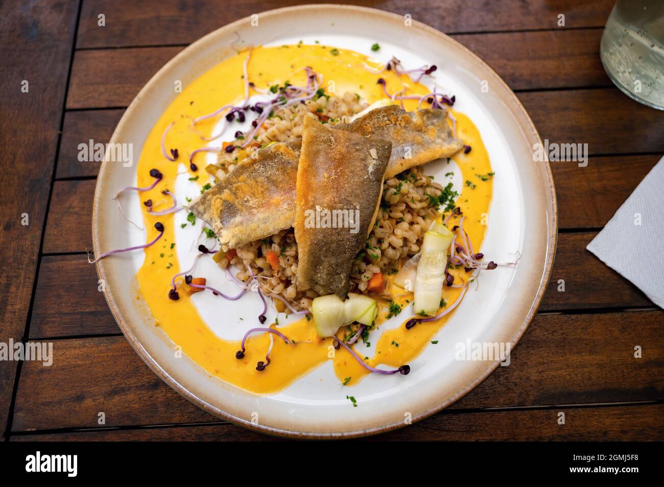 Filetto di pesce zander con semola d'orzo, zucchine, carote, germogli e salsa di senape su piatto su tavola di legno, all'aperto, closeup. Foto Stock