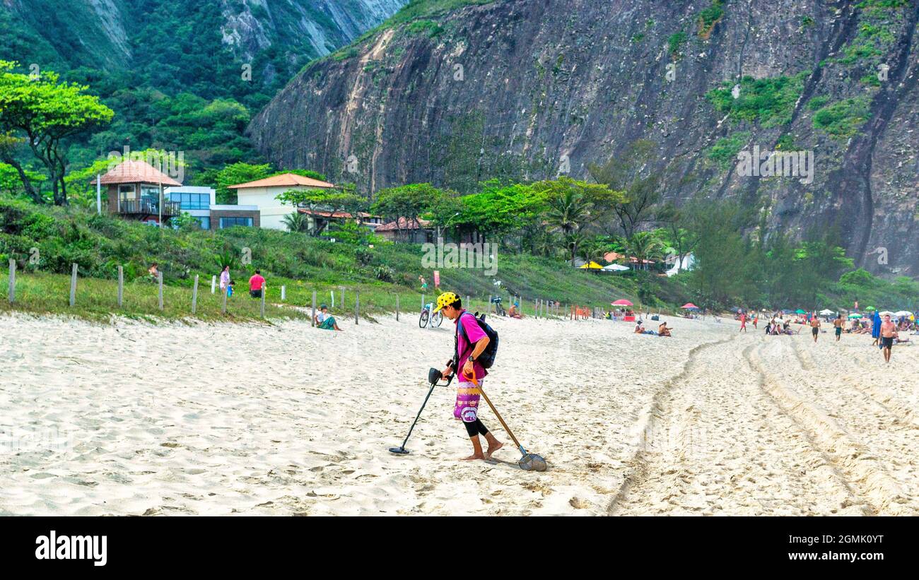 Un uomo brasiliano alla ricerca di oggetti metallici perduti, mentre i turisti godono la spiaggia di Itacoatiara a Nitreoi, Rio de Janeiro, Brasile. Con una bellissima lan Foto Stock