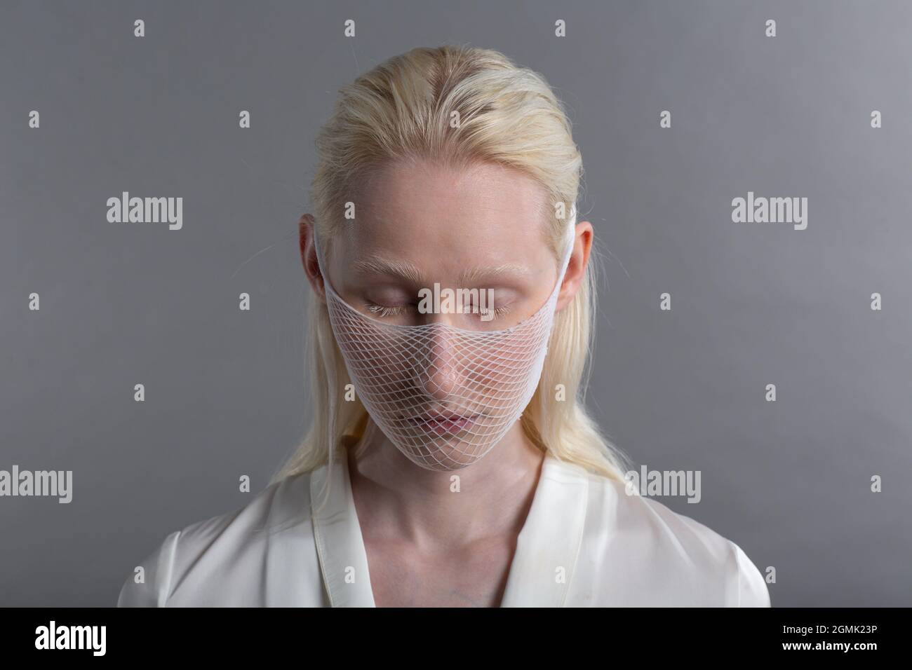 Ritratto closeup di bianco albino caucasico biondo donna moda modello indossare quarantena medico maschera viso grossolano rete a rete su sfondo grigio. Copertura del viso Foto Stock
