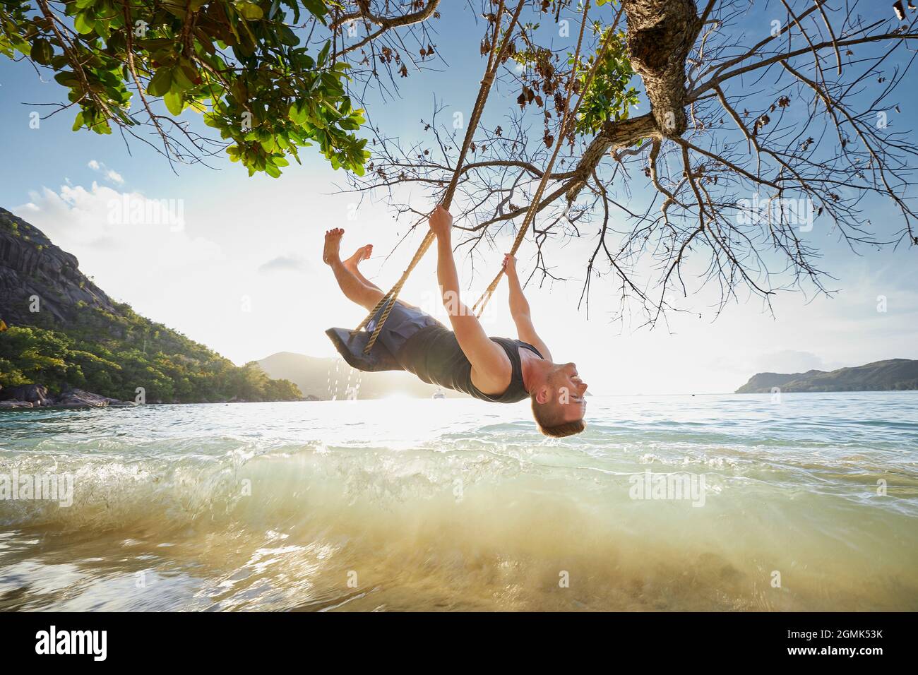 L'uomo felice sul swing che gode la vacanza. Bellissimo tramonto sulla spiaggia alle Seychelles. Foto Stock