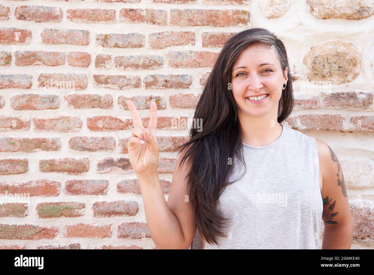 Donna sorridente con capelli lunghi che fa un segno di vittoria con la mano su un muro di mattoni. Spazio di copia per il testo Foto Stock