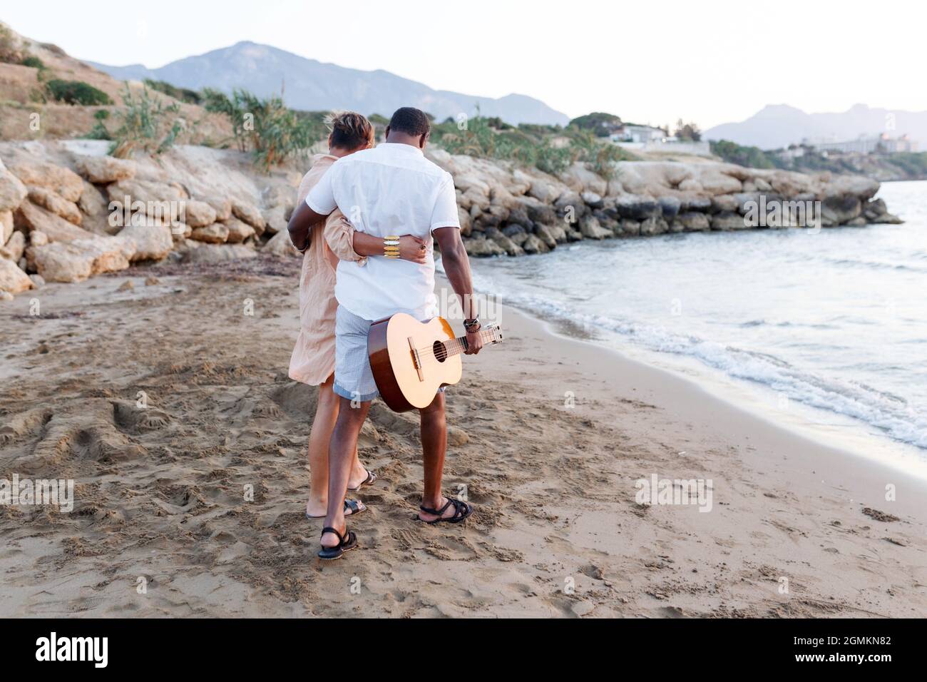Felice gara mista, coppia di mezza età abbracciando mentre si cammina sulla spiaggia tenendo una chitarra. Uomo che tiene la chitarra con la donna. Foto Stock