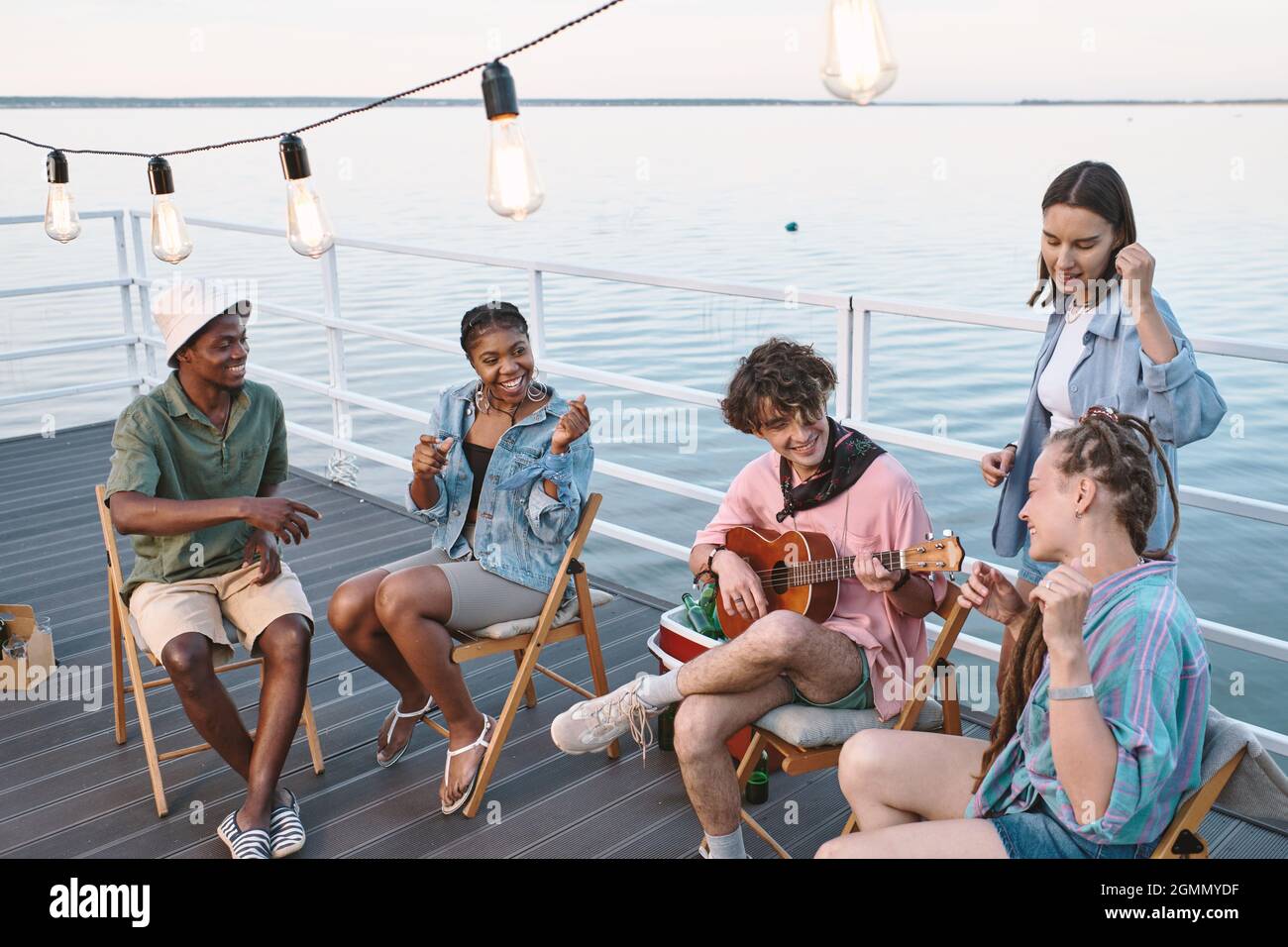 Giovani uomini e donne amichevoli cantano con la chitarra mentre trascorrono del tempo sul molo il giorno d'estate Foto Stock