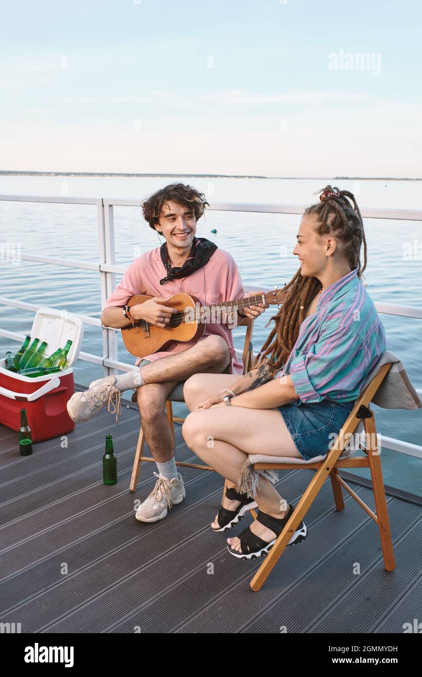 Felice giovane con chitarra guardando la sua ragazza mentre entrambi rilassarsi sul molo il giorno d'estate Foto Stock