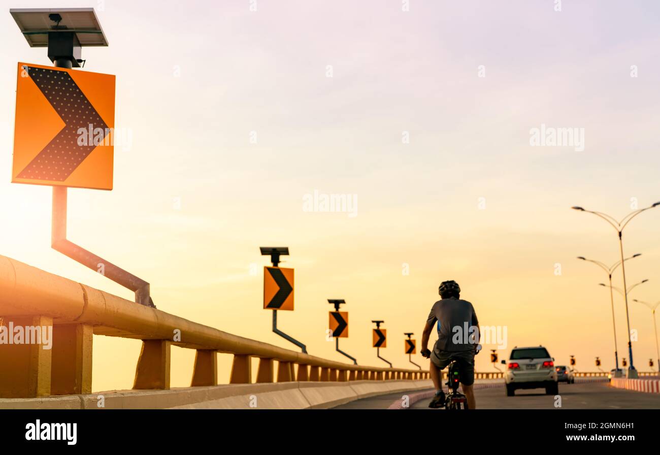 La vista posteriore dell'uomo porta il casco in bicicletta sulla strada curva dietro le auto. Guida sicura e guida su strada. Curva segnaletica stradale con solare Foto Stock