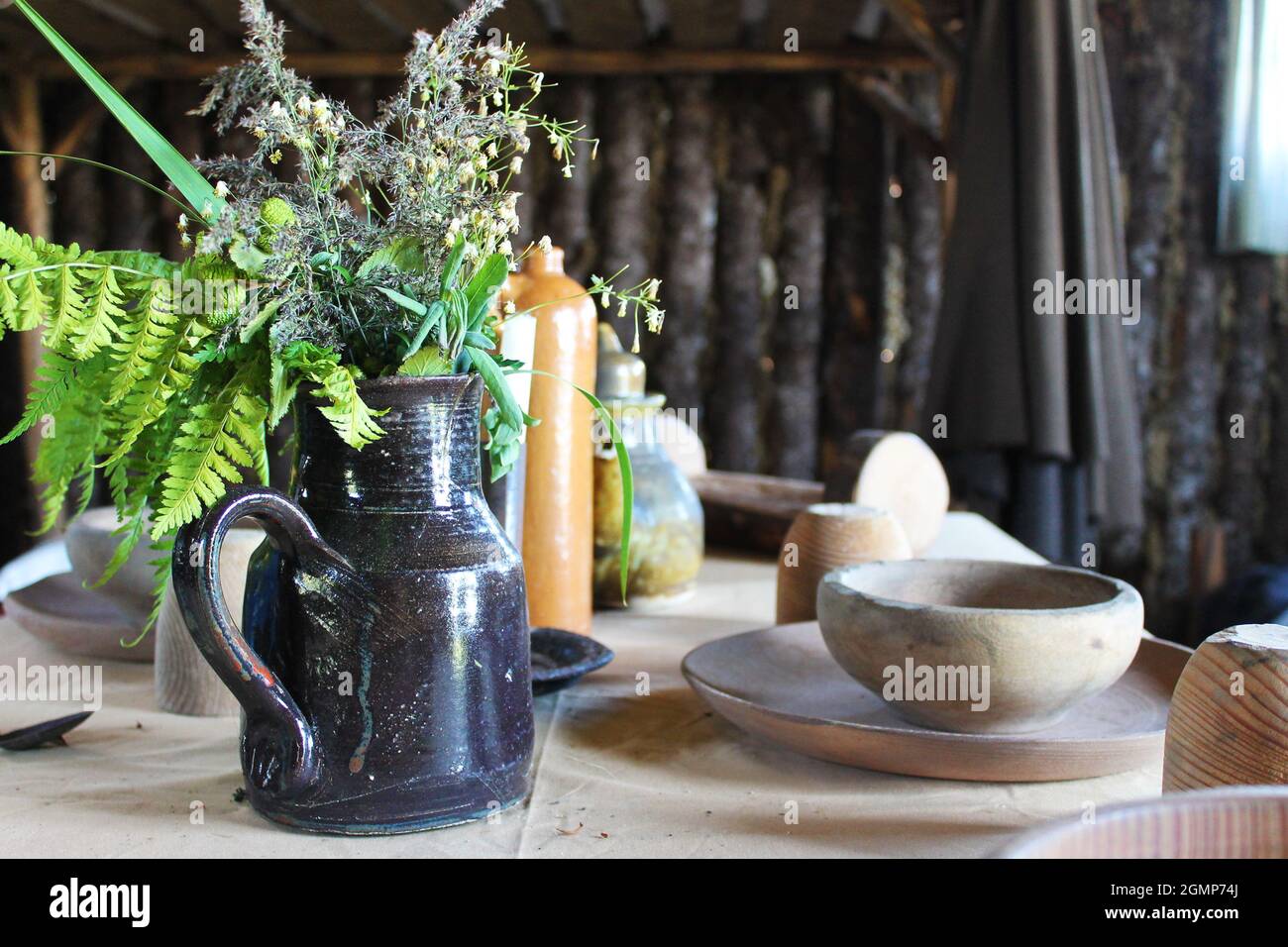 Primo piano di una caraffa in ceramica con fiori selvatici, su un tavolo con vecchio stoviglie in legno, all'interno di una cabina di tronchi. Foto Stock