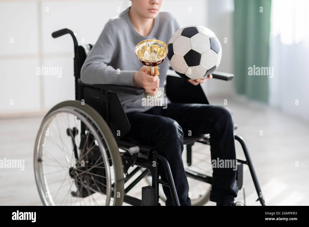 Vista ritagliata del giocatore di calcio teen in sedia a rotelle che tiene palla e trofeo, sentendosi stressato sopra il suo infortunio a casa Foto Stock