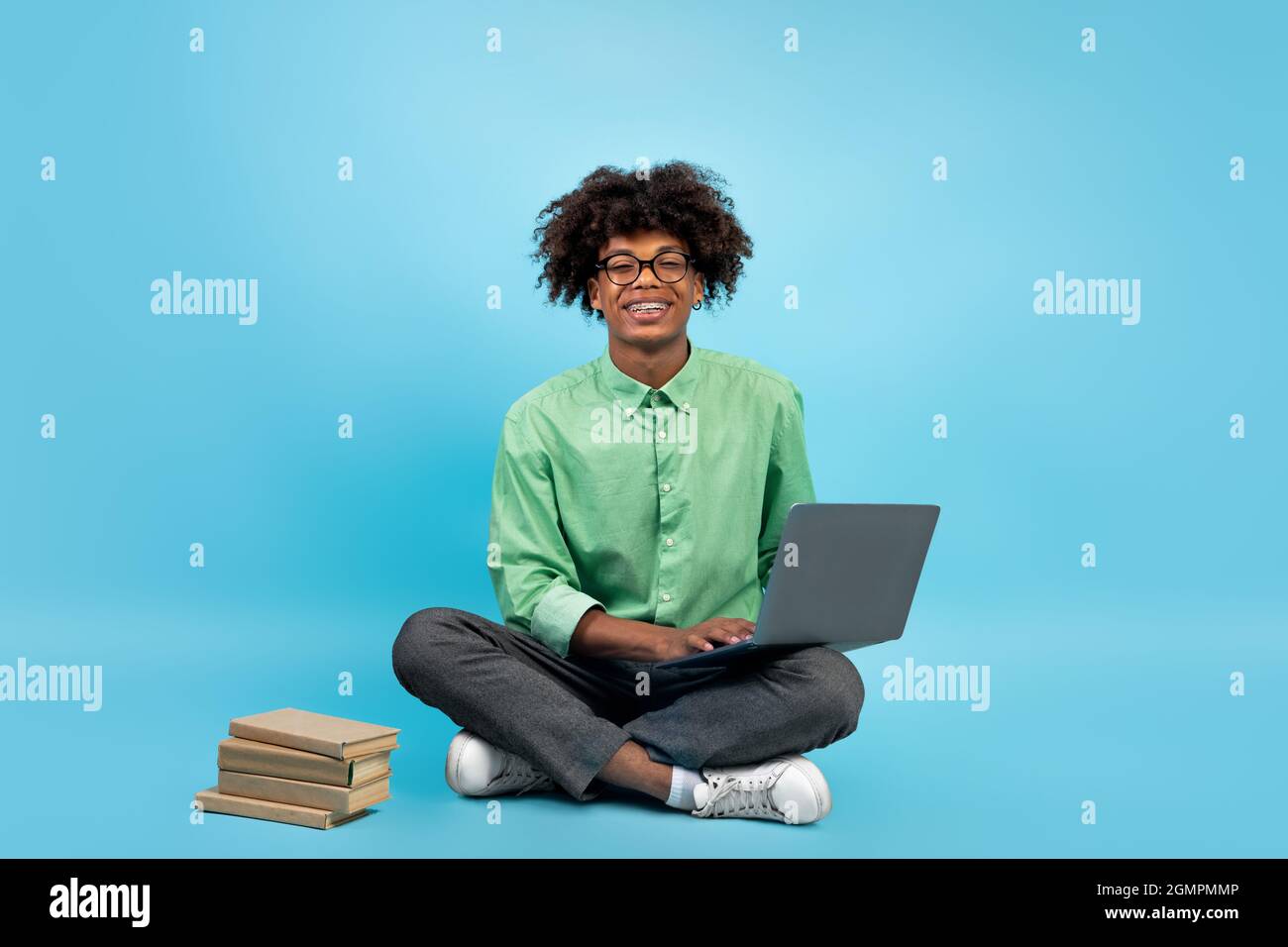 Adolescente nero eccitato con notebook e libri nelle vicinanze, seduta sul pavimento su sfondo blu studio, spazio libero Foto Stock