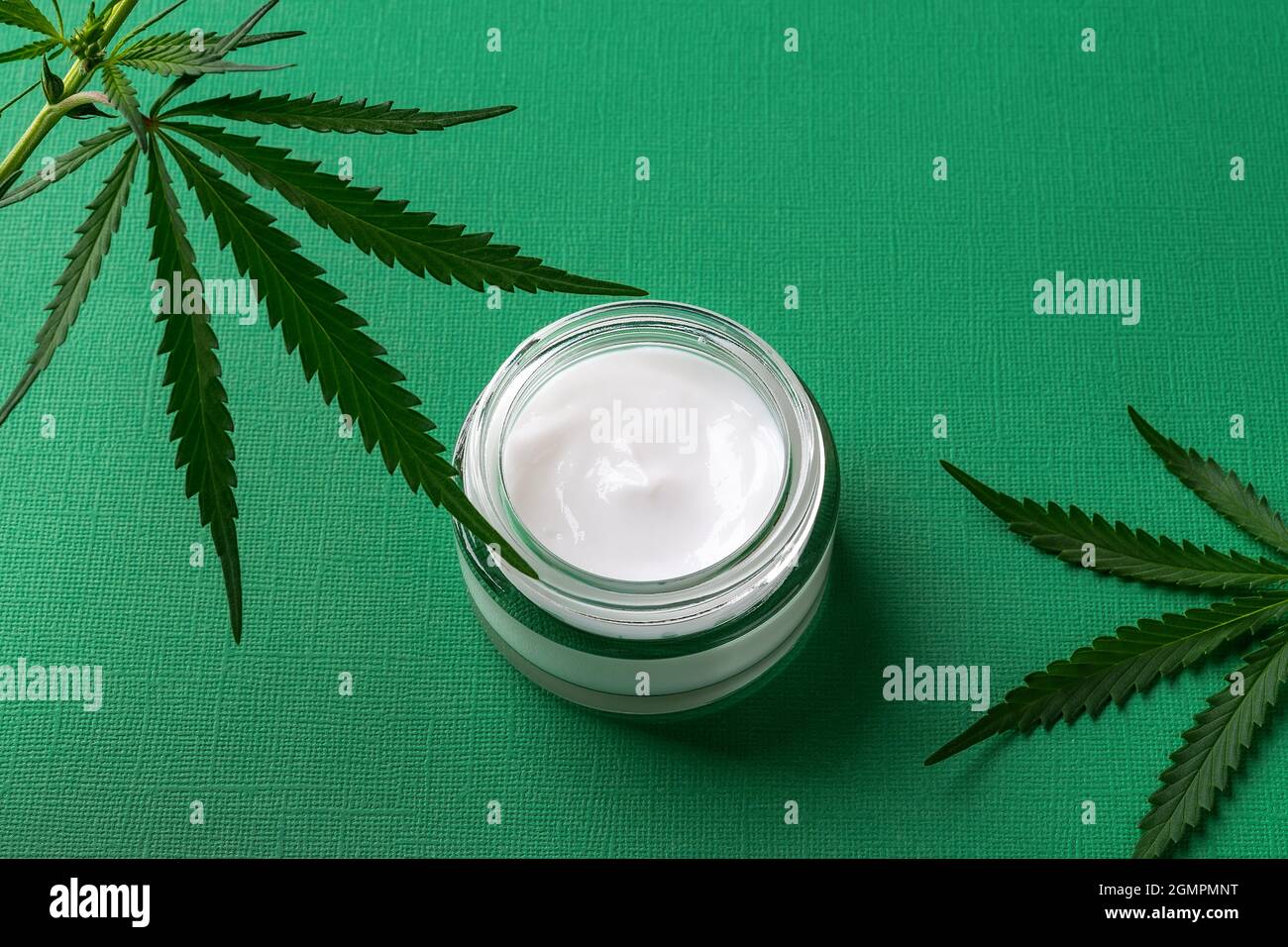 Crema viso idratante con olio cbd in vaso di vetro e foglie di cannabis sativa verde su sfondo verde testurizzato. Cosmetici anti-invecchiamento della canapa. Foto Stock