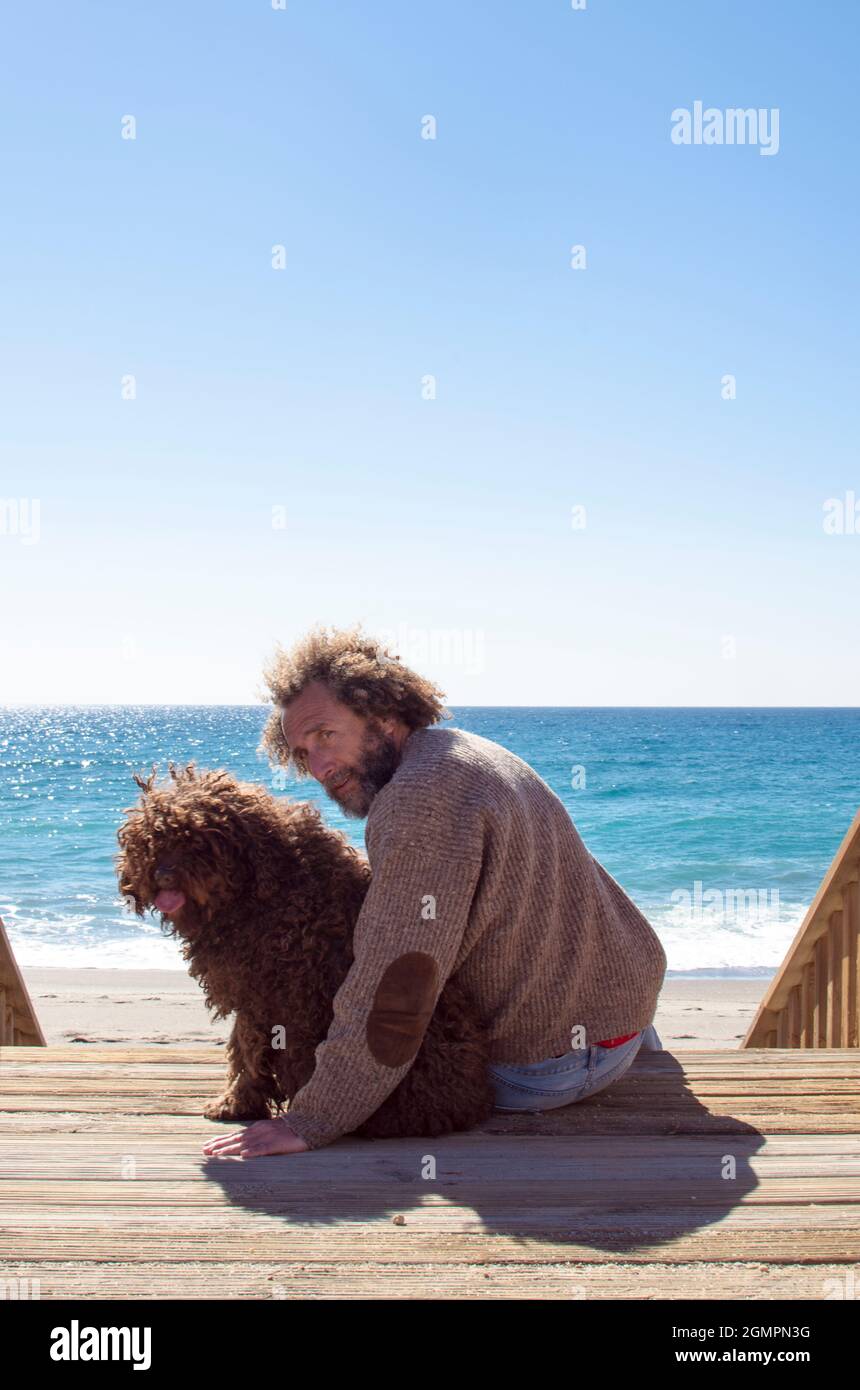 l'uomo seduto e il cane si girano verso l'alto per guardare la macchina fotografica, il mare è sullo sfondo Foto Stock