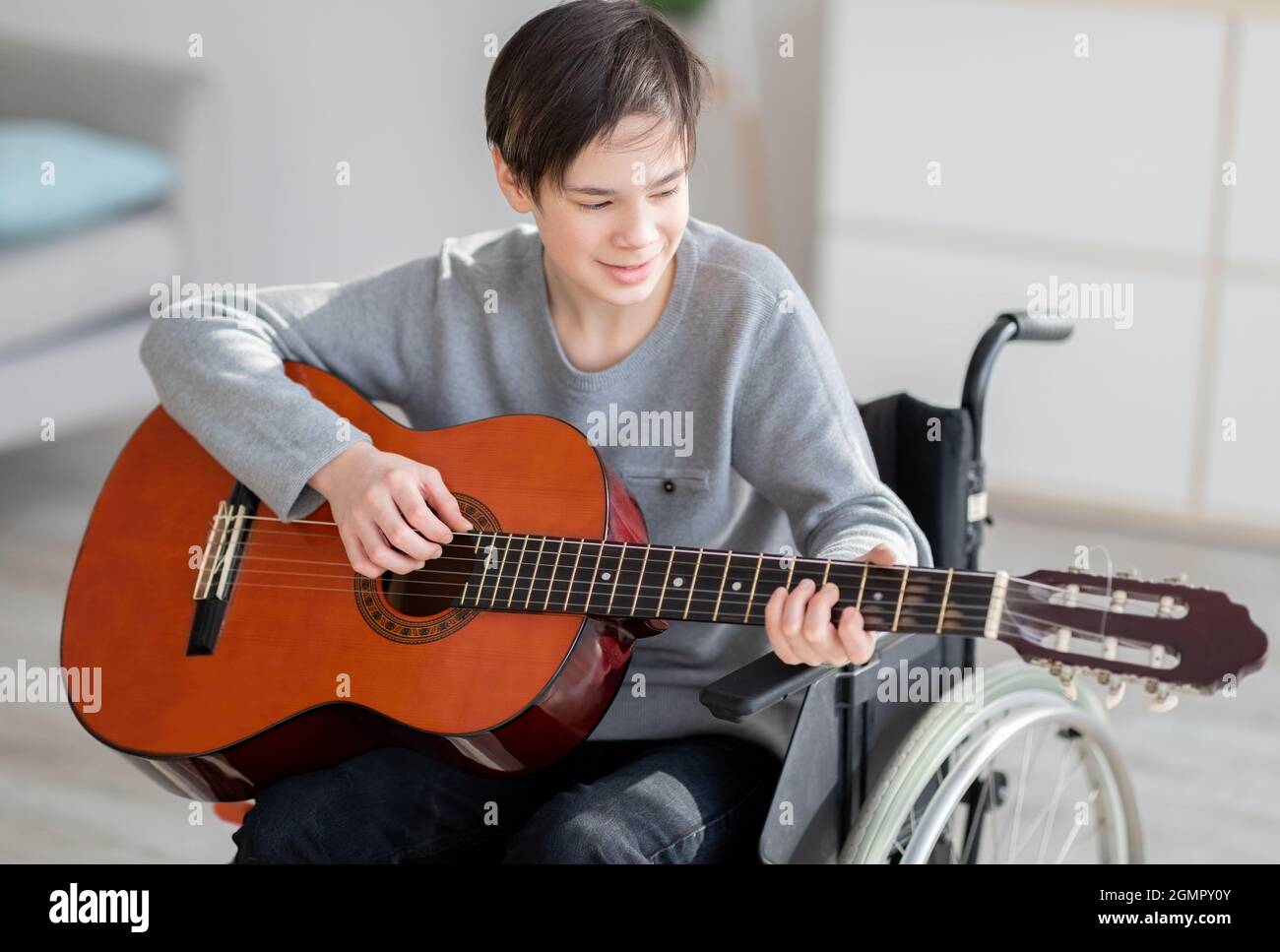 Ragazzo adolescente con disabilità allegra in sedia a rotelle imparando a suonare la chitarra e cantare al chiuso Foto Stock