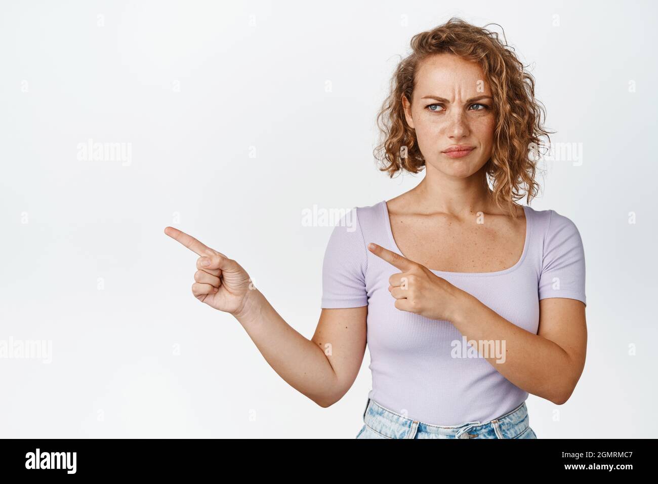 Delusa giovane donna con capelli ricci corti, accigliato e sollacciato, puntando le dita a sinistra con volto dissimile, sfondo bianco Foto Stock