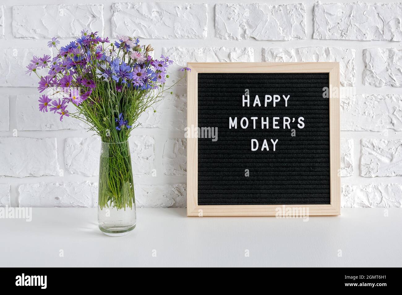 Testo del giorno delle madri felici sul bordo nero della lettera e fiori blu del bouquet in vaso sul tavolo contro la parete bianca del mattone. Giornata delle madri del concetto. Modello per gre Foto Stock