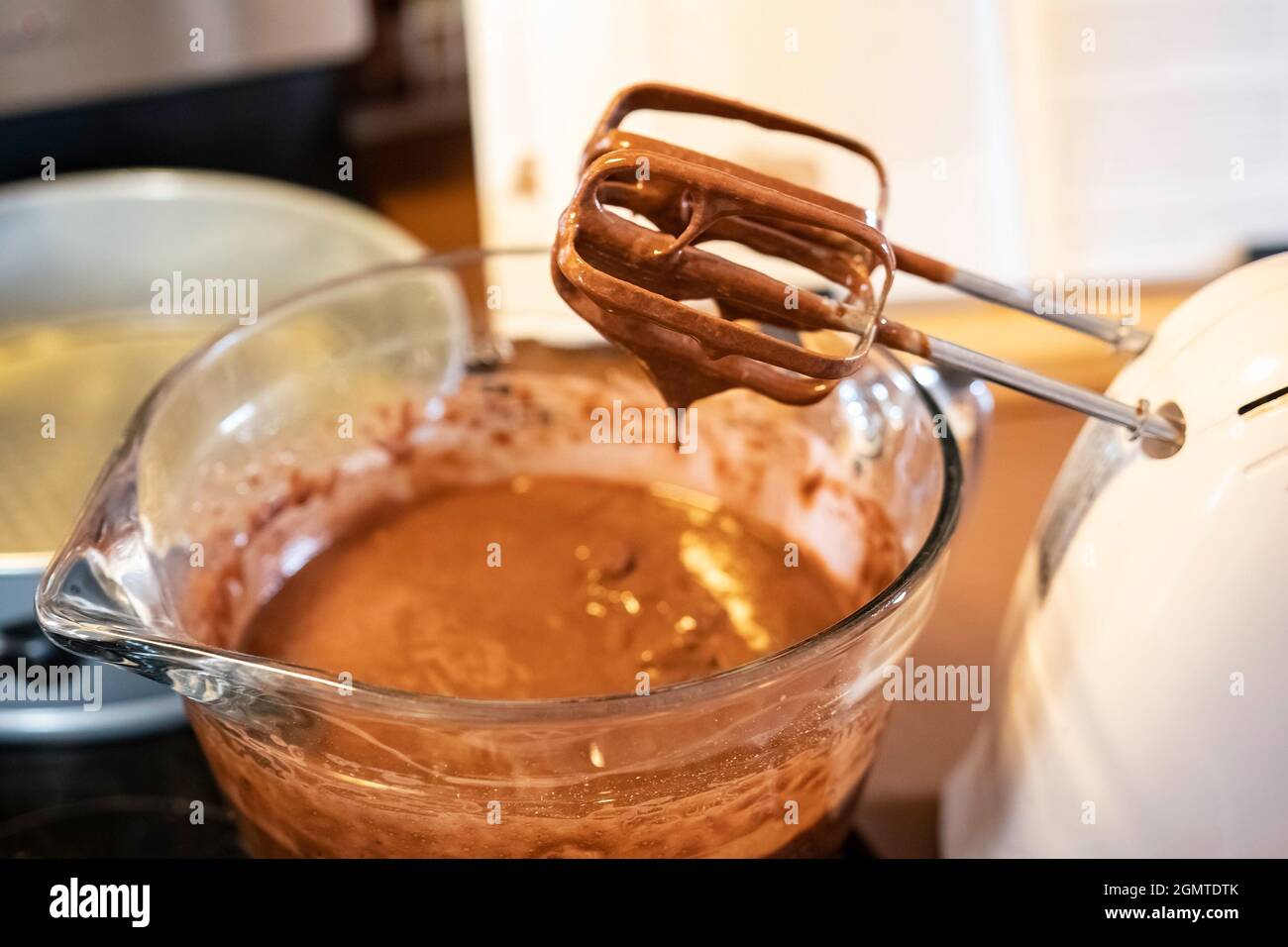 Pastella per torta al cioccolato che fuoriesce dal mixer manuale in un recipiente per la miscelazione del vetro con teglia da forno e libro di cucina in background fuori fuoco. Foto Stock