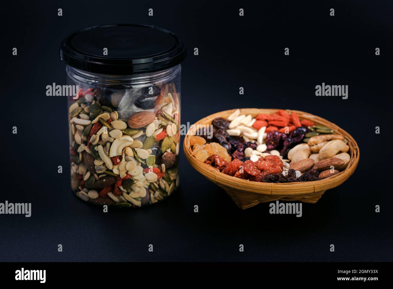Grani interi e frutta secca in un piatto di bambù e bottiglia di plastica su sfondo nero. Foto Stock