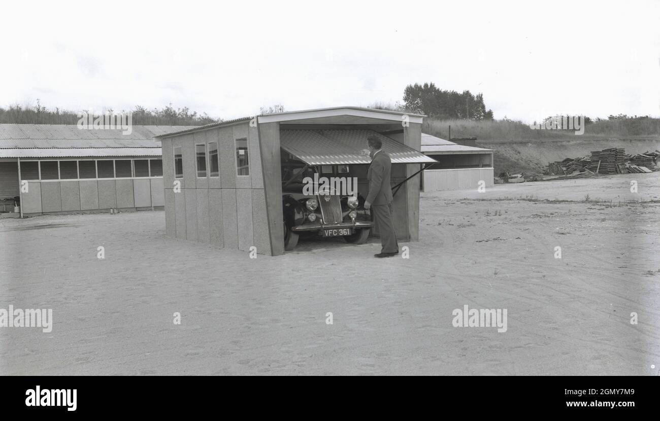 Anni '50, storico, all'esterno su un'ampia area pianeggiante in una zona industriale leggera, un uomo in un vestito che solleva la porta di roll-up di garage prefabbricato, costruito lì per scopi dimostrativi, Witney, Oxford, Inghilterra, Regno Unito. Questi garage o ripari per motori sono stati realizzati con pannelli in calcestruzzo prefabbricato imbullonati insieme in sezioni, con un tetto in metallo ondulato e metallo arrotolarsi o su e sopra la porta. Con l'aumento della proprietà delle auto negli anni '50 e '60, questi garage auto-costruiti o kit in cemento sono diventati la cosa da avere e un gran numero di aziende hanno offerto tali garage fai-da-te al 'casalingo pratico'. Foto Stock