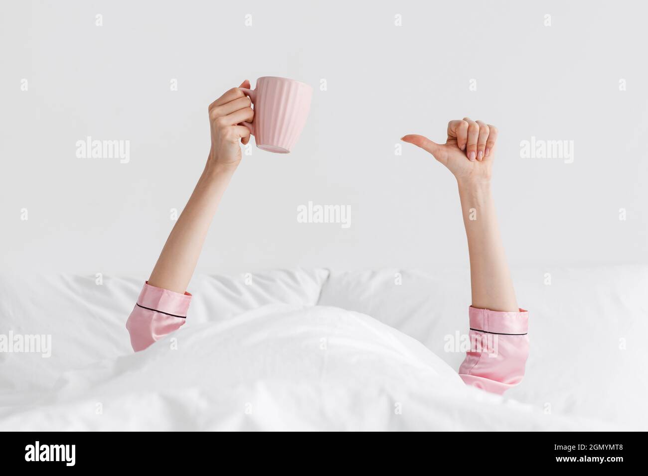 Mani di giovane bionda europea in pajama rosa sporgono dalla coperta bianca e tengono una tazza di gustoso caffè Foto Stock